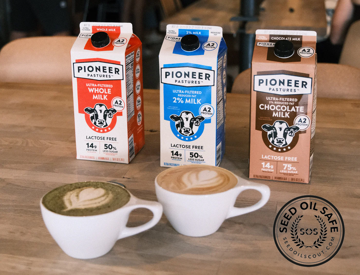 Three cartons of Pioneer Pastures milk on a wooden table with two cups of coffee.