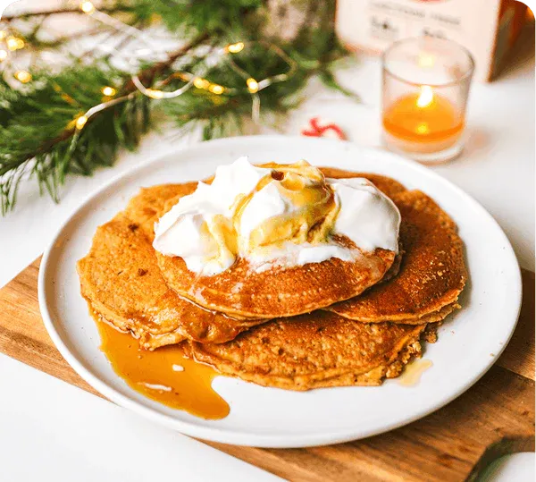 Stack of pancakes with syrup and whipped cream on a plate, set against a festive background with lights and a candle.