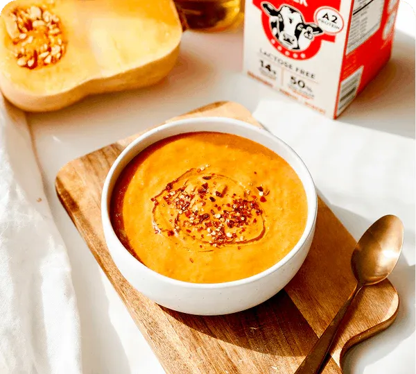 Bowl of pumpkin soup on a wooden board with a butternut squash and a carton of milk in the background.
